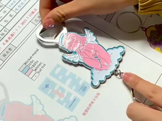 A hand holding a custom-shaped enamel pin featuring an angel on a cloud with pink and light blue details, integrated with a bottle opener top and a keychain ring attached at the bottom.