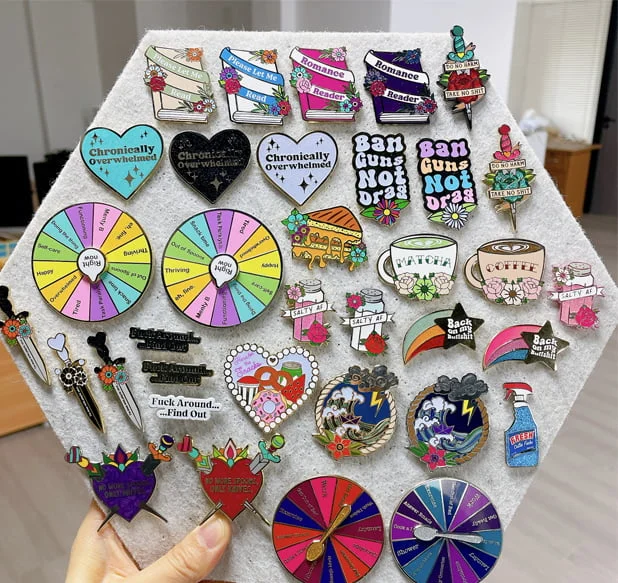 A collection of various custom enamel pins displayed on a hexagonal board, featuring book designs, heart shapes with text, spinner wheels, coffee cups, daggers, and colorful thematic artwork.
