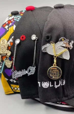Three baseball caps decorated with high quality custom enamel pin hat clips featuring dangling chains, rhinestones, and metallic designs.