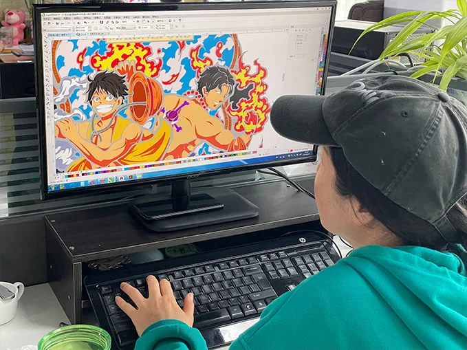 SYD designer is working at a computer, with design software open on the screen displaying a colorful digital illustration of anime characters Luffy and Ace for a custom enamel pin.