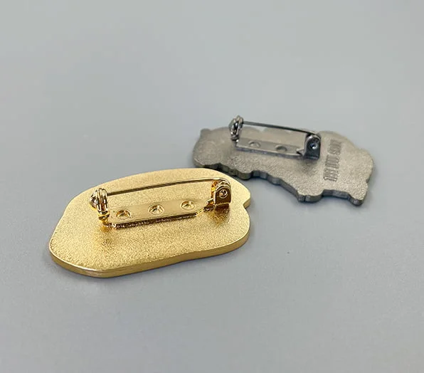 Two custom pins shown from the back, featuring gold-toned and silver-toned safety pin backings securely fastened to textured metal plating.