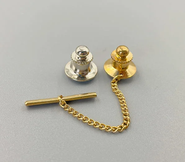 A gold-toned tie tack pin backing featuring a clutch, a short chain, and a horizontal bar pin, alongside a silver-toned butterfly clutch.