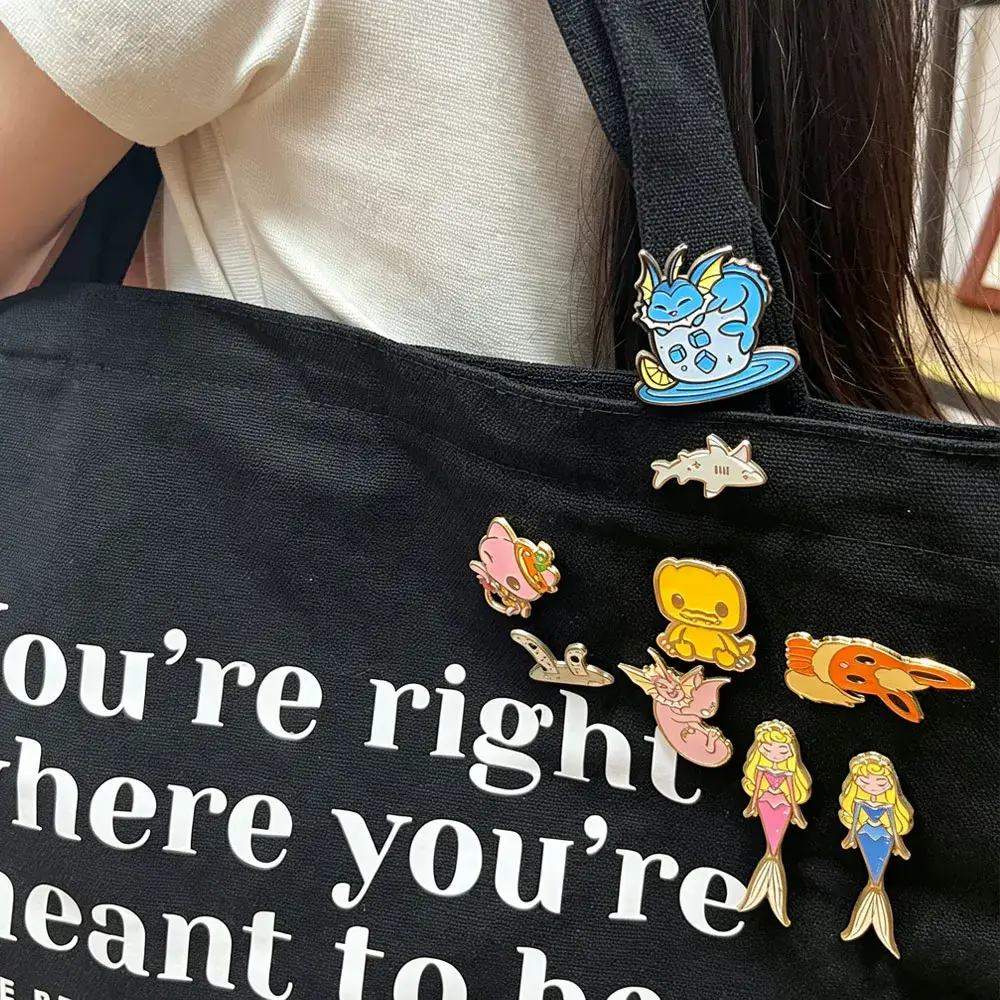 Collection of enamel pins featuring various cute creatures (dragons, mermaids, animals) displayed on a black canvas tote bag.