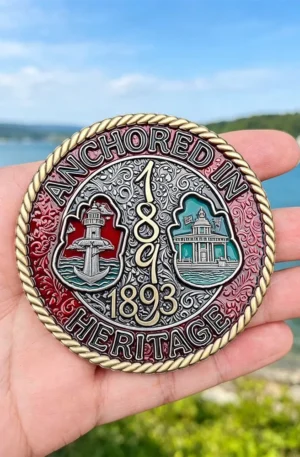 Custom challenge coin with a rope-edge border featuring "Anchored in Heritage" and "1893" text, a lighthouse motif, and colorful enamel accents held against an outdoor waterfront background.