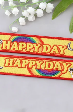 Custom embroidered keychains featuring bright yellow fabric tags with red "HAPPY DAY" lettering, colorful rainbow and crescent moon motifs, red merrowed borders, and metal split keyrings.
