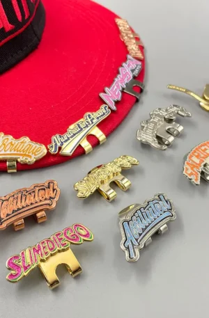 Custom Enamel Hat Clip - Collection of "AABA Boutique," "Slimediego," "Affiliated," and "Sacrifices" Magnetic Hat Clips Displayed Alongside a Red Baseball Cap