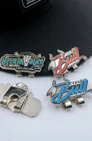Custom Enamel Hat Clip - Assorted "Gfour Hat Co." and "All Bull No Shit" Magnetic Hat Clips Showing Front Designs and Back Clip Mechanism Placed on a Black Baseball Cap