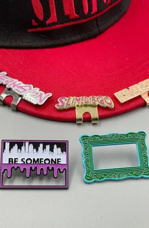 Custom Enamel Hat Clips and Lapel Pins - Assorted "Neonsan Tokyo," "Slimediego," and "Houston" Magnetic Hat Clips Attached to a Red Baseball Cap, Accompanied by "Be Someone" City Skyline Pin and an Ornate Green Picture Frame Pin