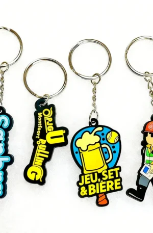 Custom PVC keychain set featuring four flexible rubber designs with split keyrings and link chains: a blue Sanbra text tag, a black and yellow OPJG Pulling logo, a beer and tennis racquet motif, and a cartoon construction worker figure.