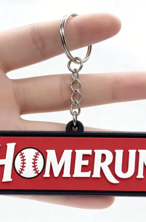 Hand holding a custom PVC keychain featuring a red rectangular tag with white "HOMERUN" lettering where the letter O is a baseball with red stitching, surrounded by a black border, link chain, and split keyring.