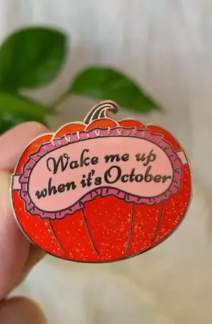 Custom enamel pin featuring a sparkly red pumpkin with a pink sleep mask graphic and the text "Wake me up when it's October" with gold plating