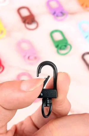 Hand holding a black metal snap hook showcasing its spring-loaded gate and swivel ring, surrounded by a blurry assortment of colorful snap hooks in the background.